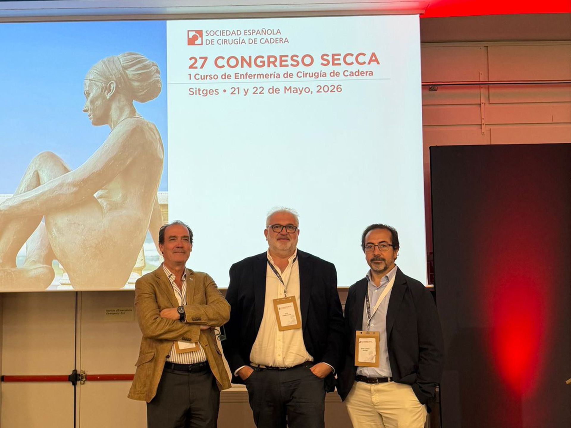 El Doctor Boris García posando junto a otros dos especialistas médicos frente a la pantalla de presentación del 27º Congreso de la Sociedad Española de Cirugía de Cadera (SECCA) celebrado en Sitges.